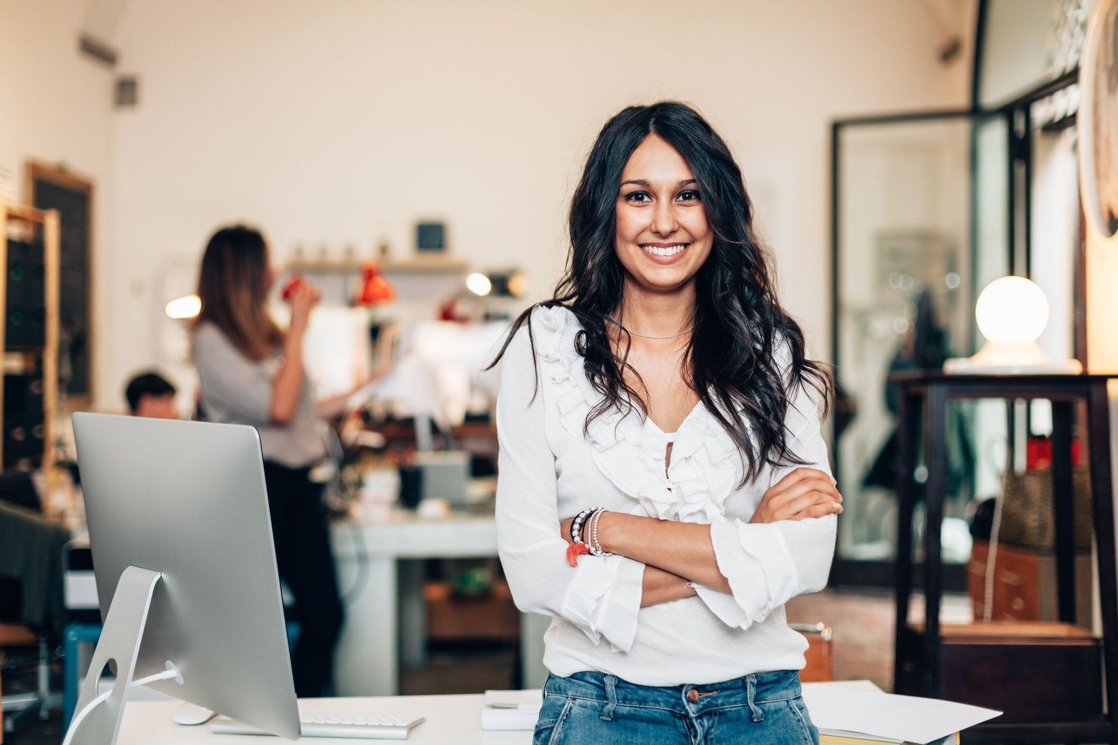 A woman is standing in front of a computer in an office with her arms crossed.
