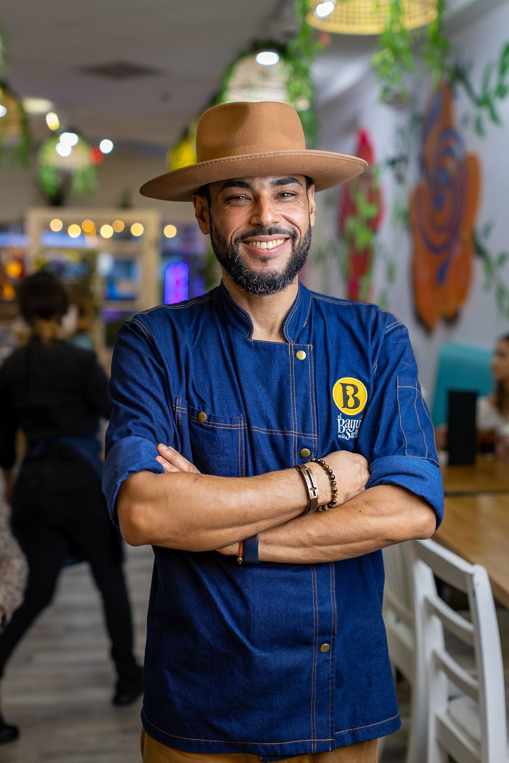 Cheff Ayala wearing a hat and a chef 's jacket is standing in a restaurant with his arms crossed .