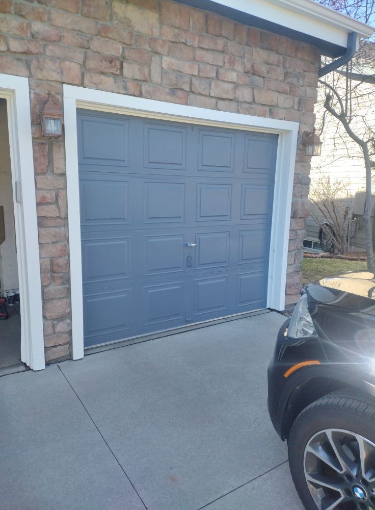 A gray, multi-panel garage door set in a stone-faced exterior wall, with a partial view of a car parked in the driveway.