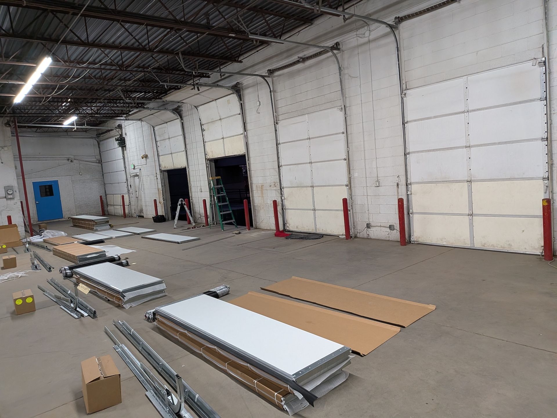 Inside a warehouse, construction materials and ladders are on the concrete floor near several closed loading dock doors.