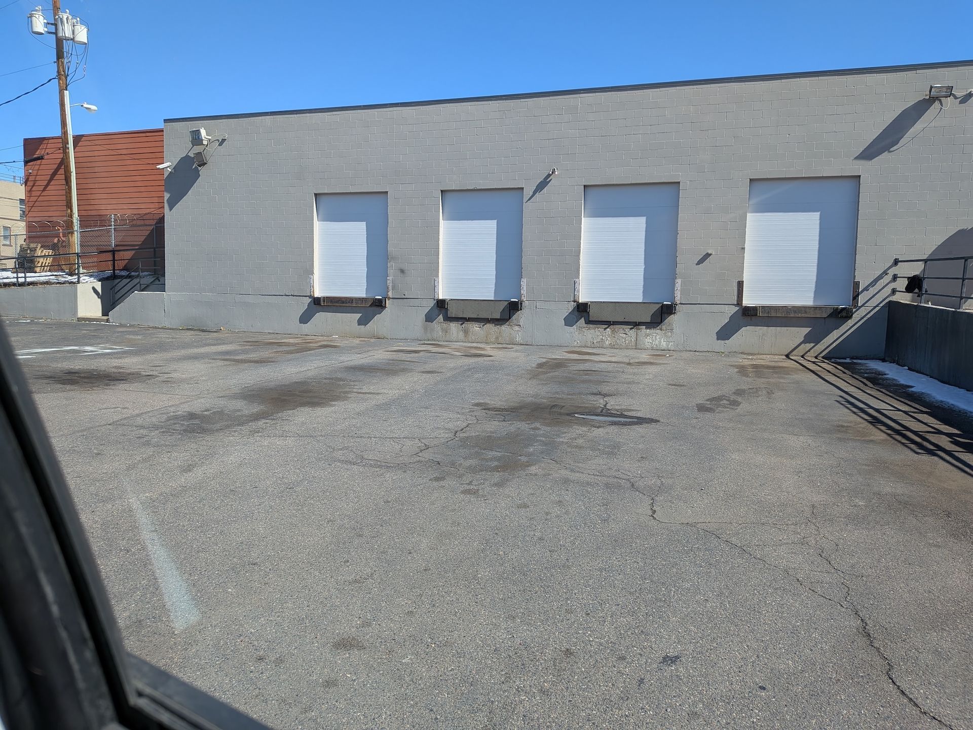 A gray brick building with four white roll-up loading dock doors above a paved parking lot on a sunny day.