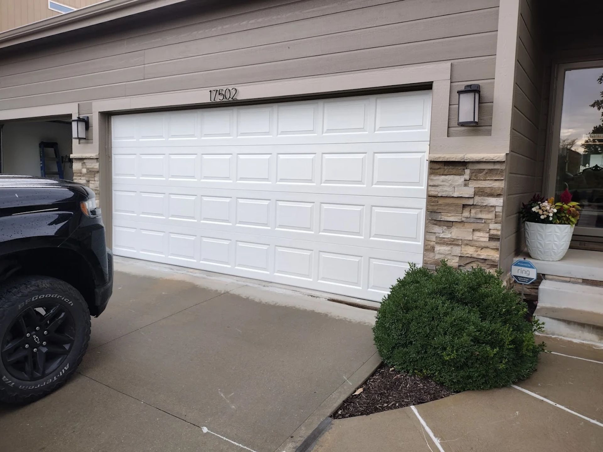 A white multi-panel garage door on a house with beige siding and stone accents, next to a black vehicle and a shrub.
