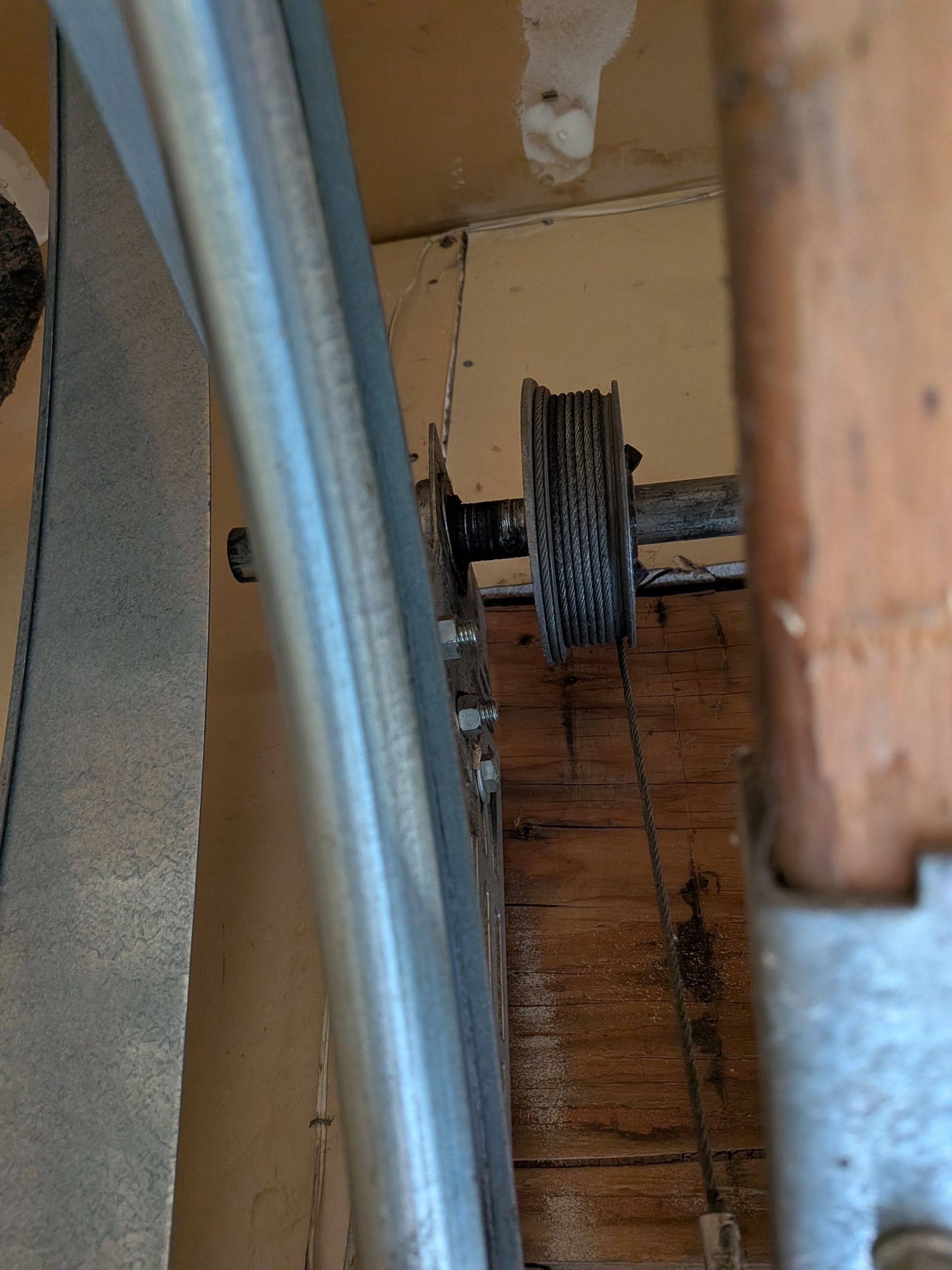 A close-up view of a metal cable wrapped around a garage door pulley drum attached to a shaft near a wooden ceiling.