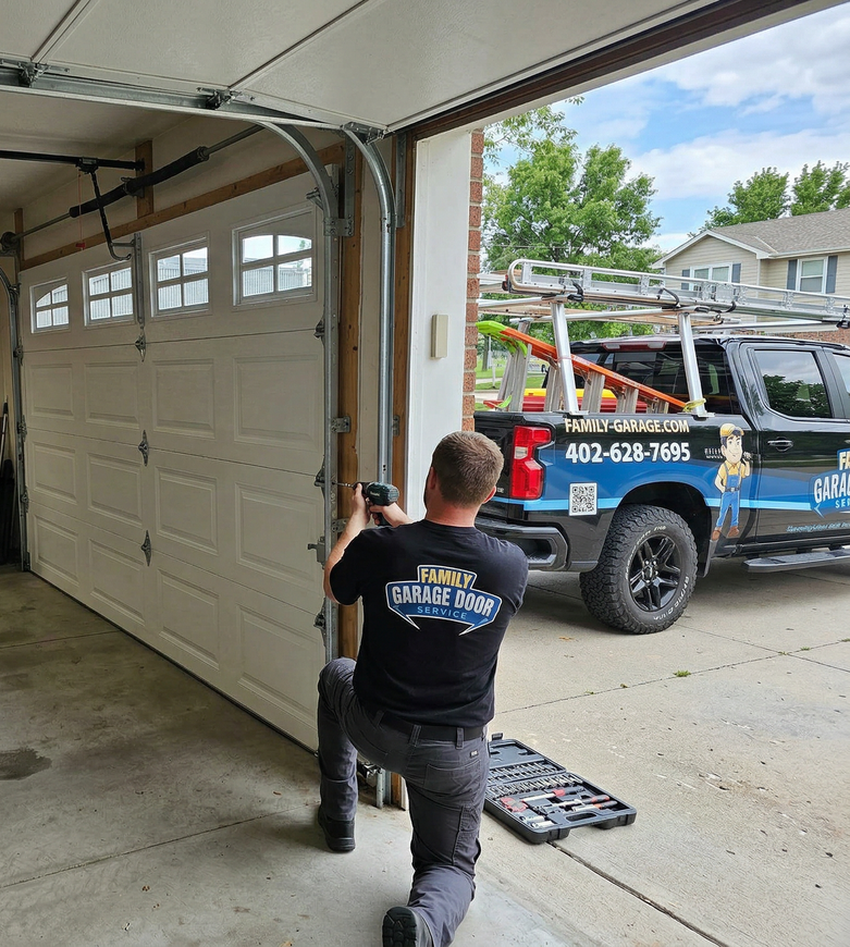 A technician in a black shirt kneels while repairing a white garage door, with a work truck parked in the driveway.