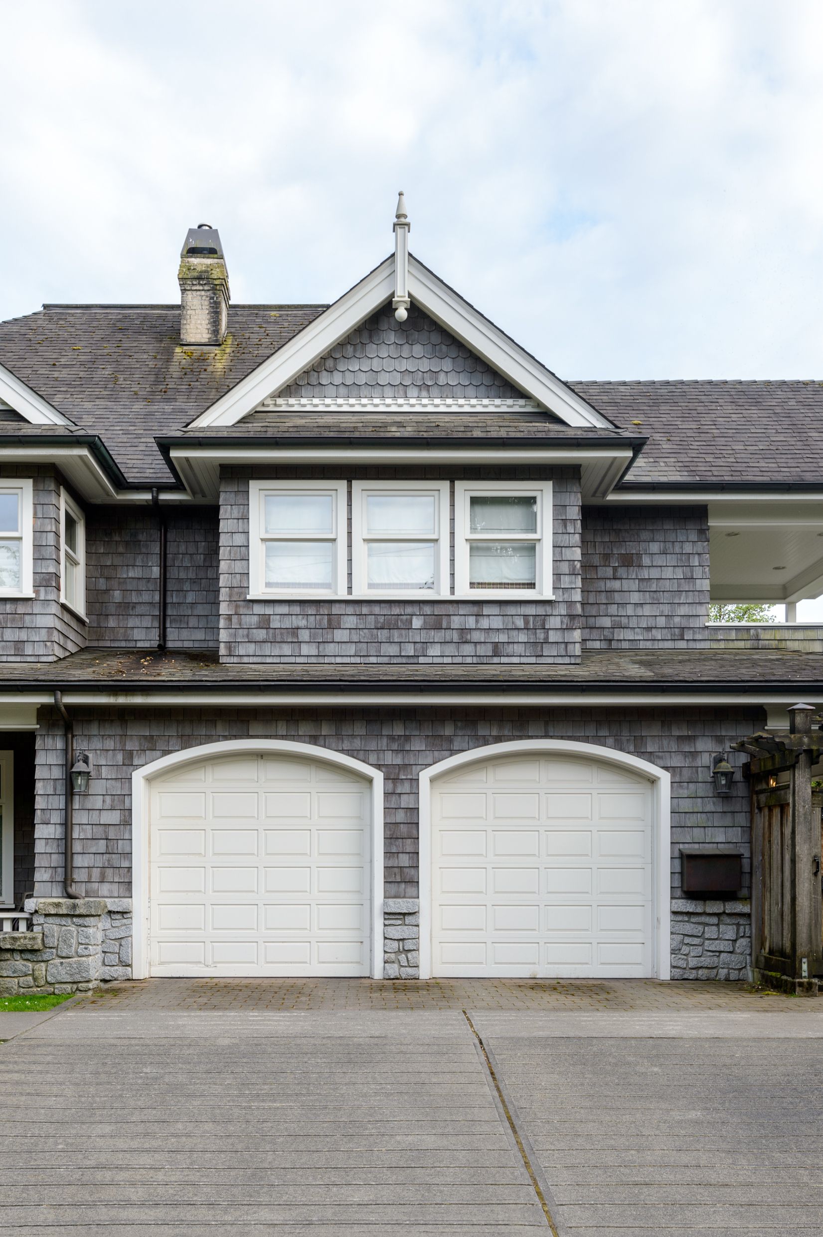 A grey two-story house with two white garage doors and a gabled roof featuring decorative shingle detailing.