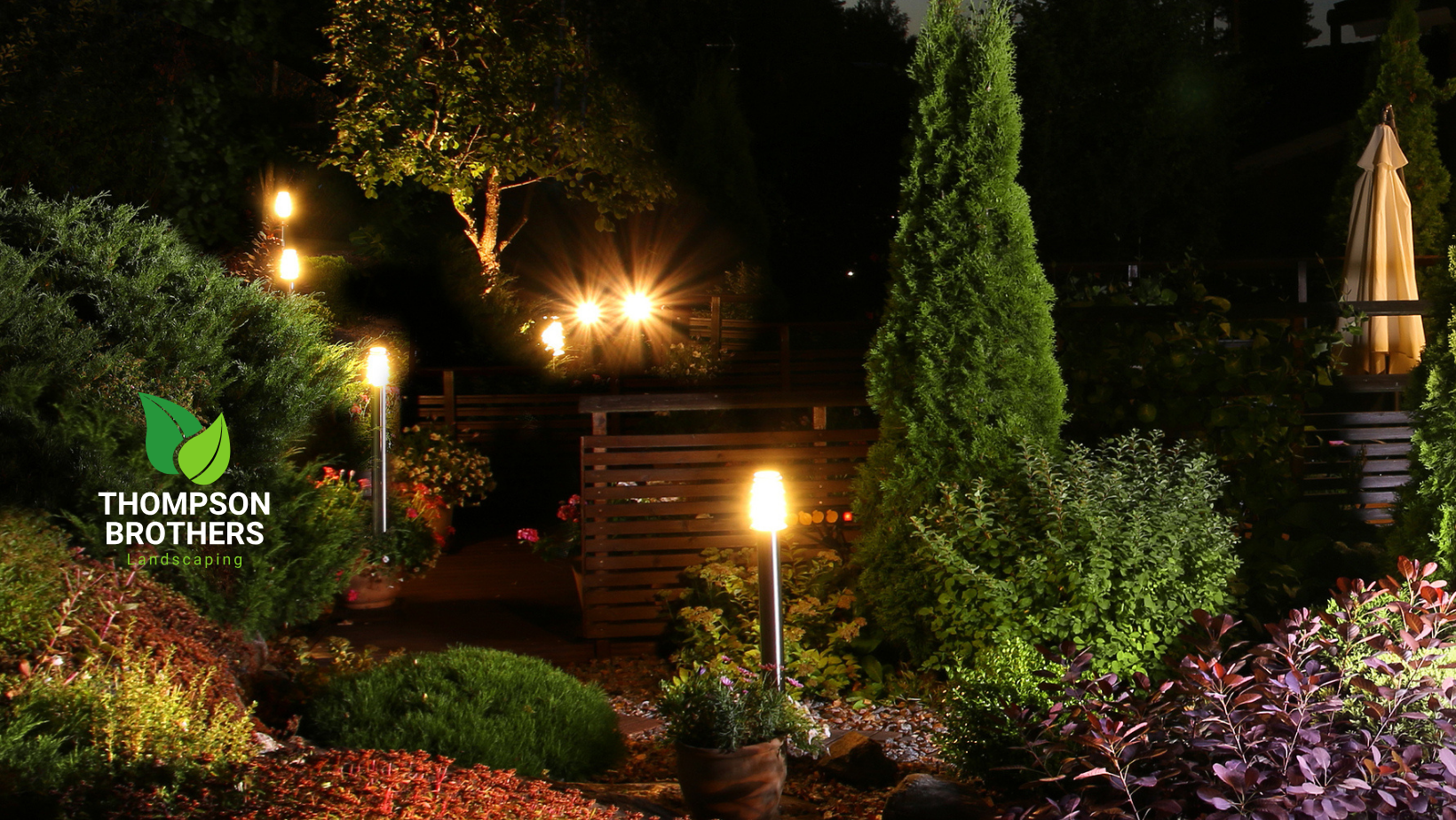 Lit backyard garden path at night with decorative lights. Bushes, trees, and wooden fence.