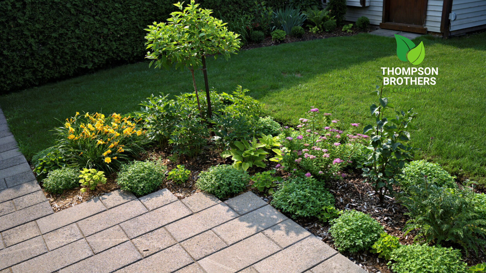 Well-manicured garden bed with various plants bordering a brick pathway and lawn.