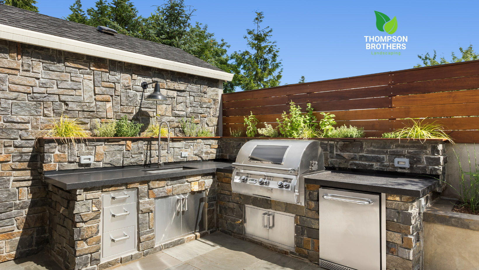 An outdoor kitchen with a grill on a paver patio and a wooden fence.
