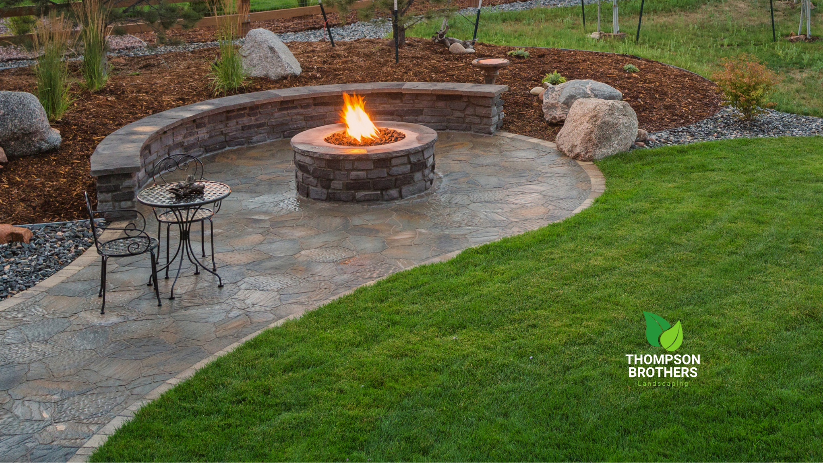 Fire pit area with curved stone wall, patio, and lawn. Fire burning in pit.