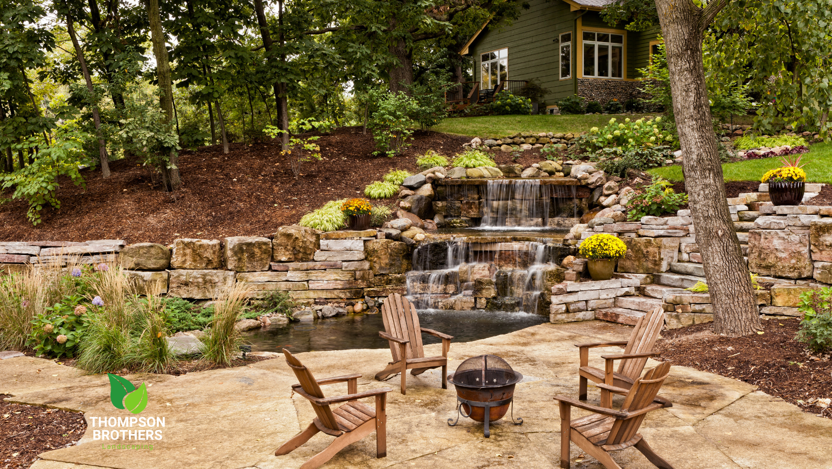 A patio with chairs and a fire pit in front of a waterfall and retaining walls.