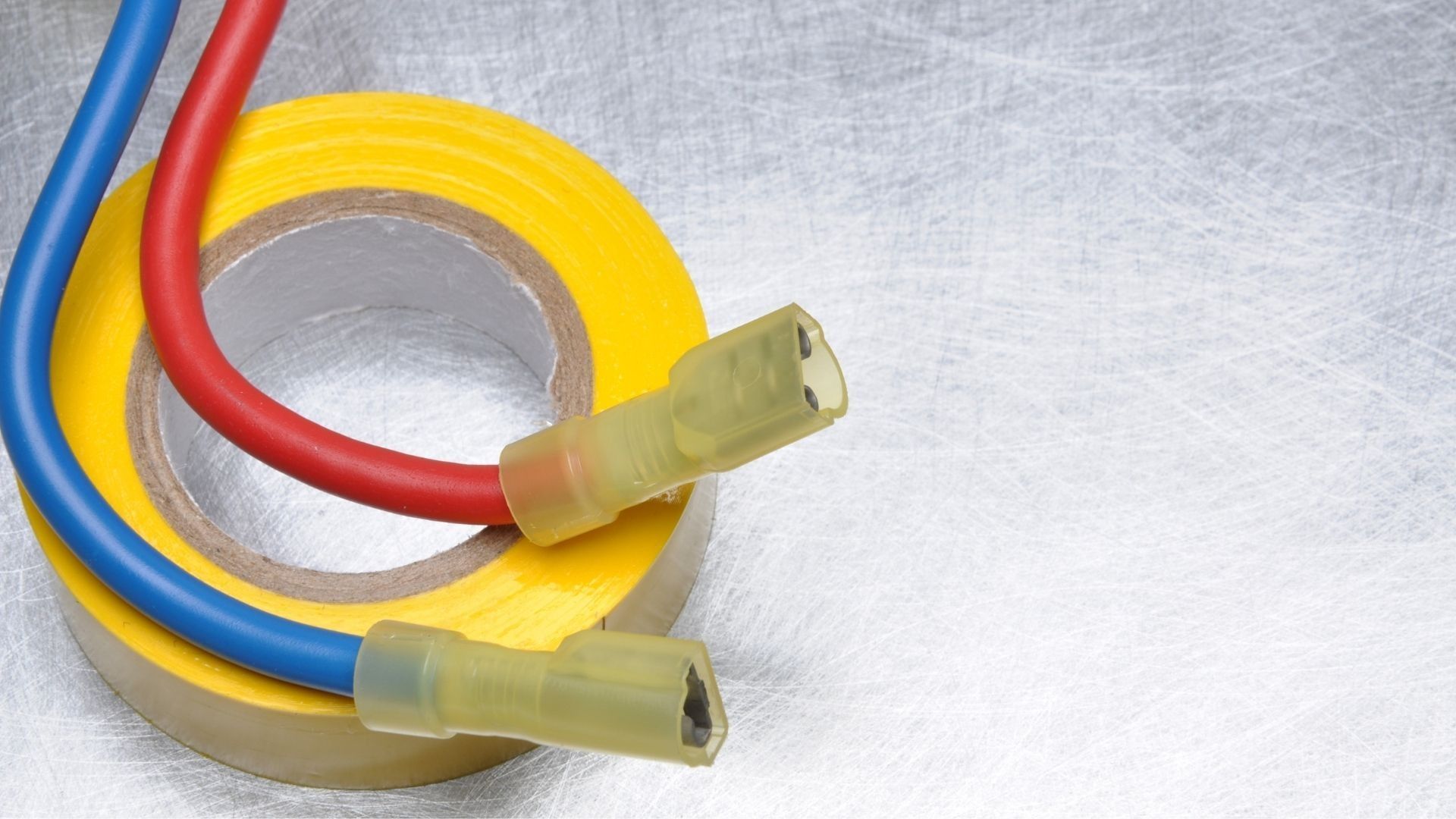 Red and blue electrical wires with yellow connectors, on a roll of yellow electrical tape, against a metallic surface.