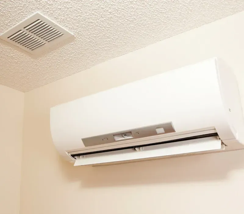 White air conditioner mounted on a light beige wall, with a ceiling vent above.