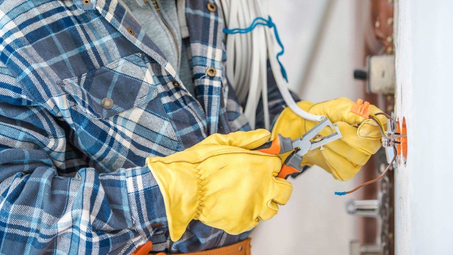 Electrician wearing yellow gloves working on electrical wiring near a wall outlet; indoors.