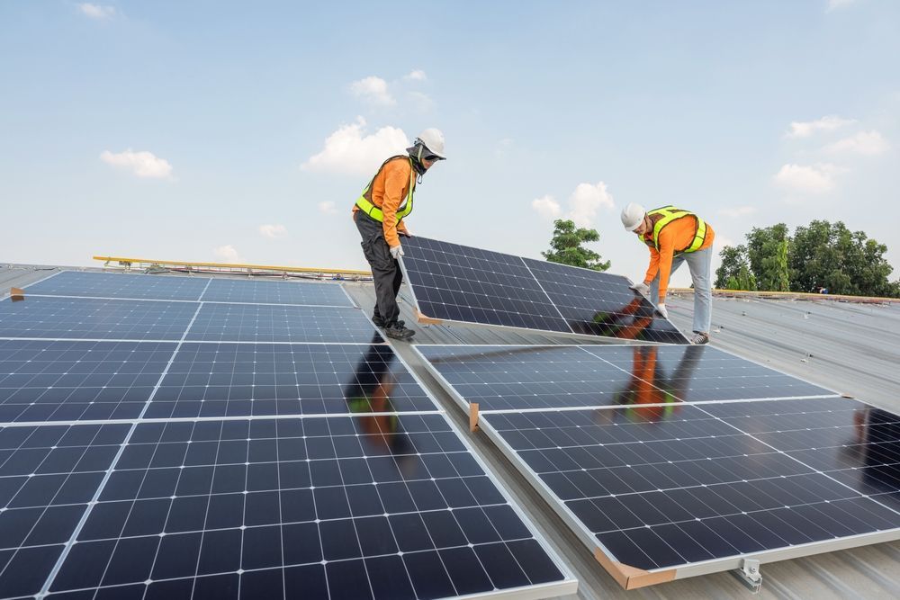 Two workers in safety vests installing a solar panel on a rooftop under a blue sky.