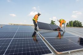 Two workers in safety vests installing a solar panel on a rooftop under a blue sky.