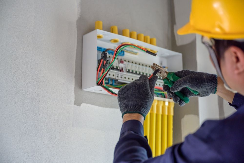 Electrician in a yellow hard hat working on a circuit breaker panel, wiring visible.