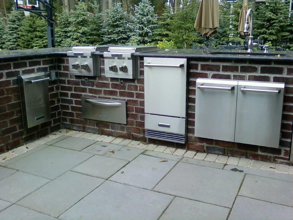 Outdoor kitchen built into a brick wall featuring stainless steel appliances, cabinets, and a sink on a stone patio.