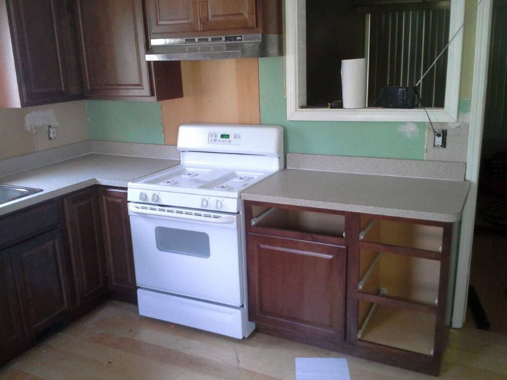 A kitchen under renovation featuring dark wood cabinets, a white stove, gray countertops, and exposed green wall areas.