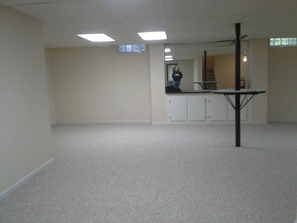 A large, beige, unfinished basement with carpet, drop ceiling lights, a mirrored wall, and a central structural pole.