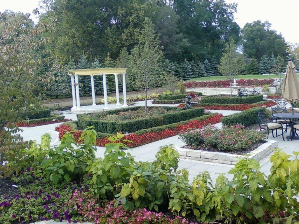 A garden features a white gazebo, geometric flower beds with red flowers, manicured hedges, and a small patio area.