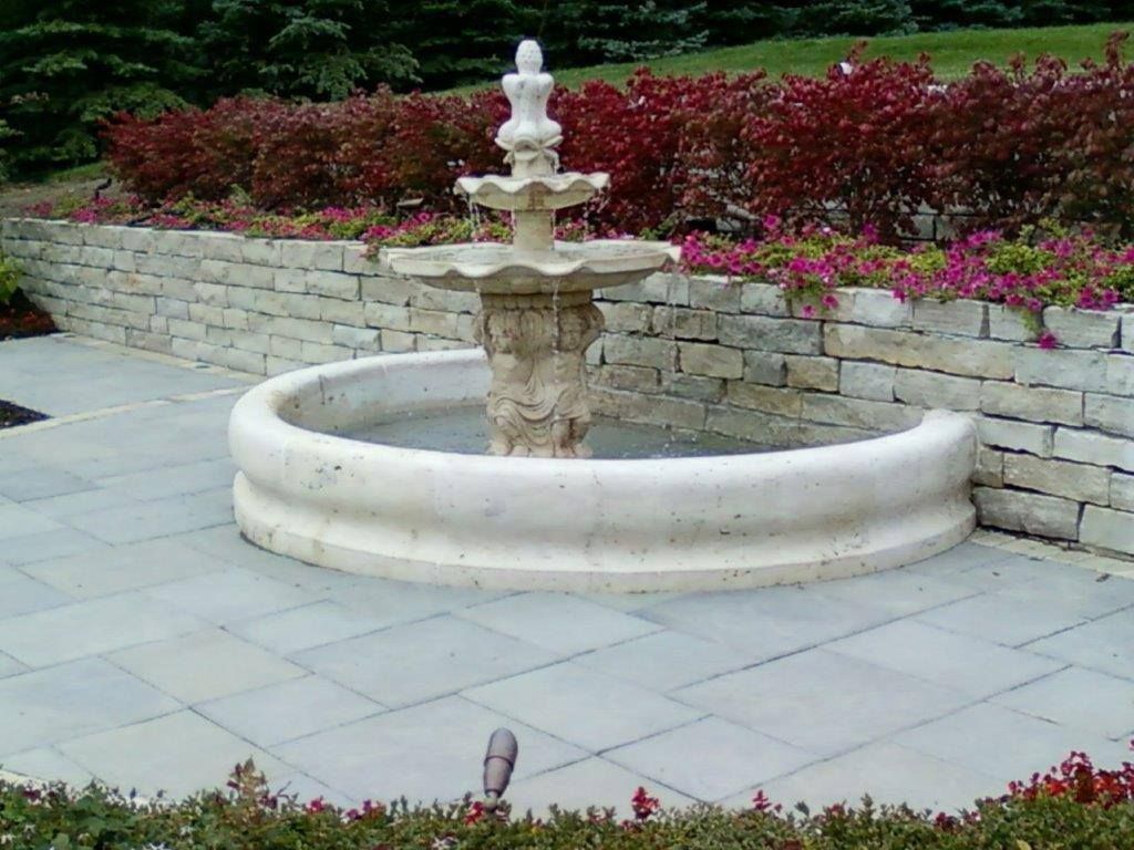A three-tiered stone fountain sits in a circular basin on a stone patio, backed by a low retaining wall and garden.
