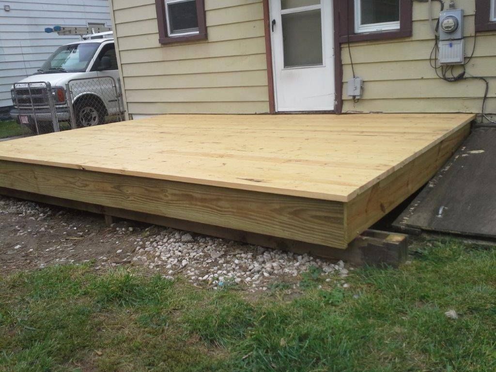 A newly constructed wooden deck platform sits attached to the side of a house next to a parked vehicle.