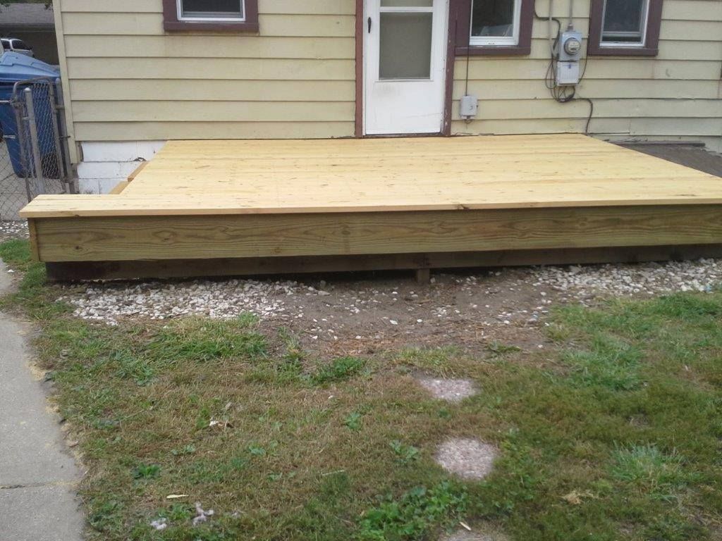 A new wooden deck attached to the back of a light-yellow house, situated above a bed of gravel in a grassy yard.