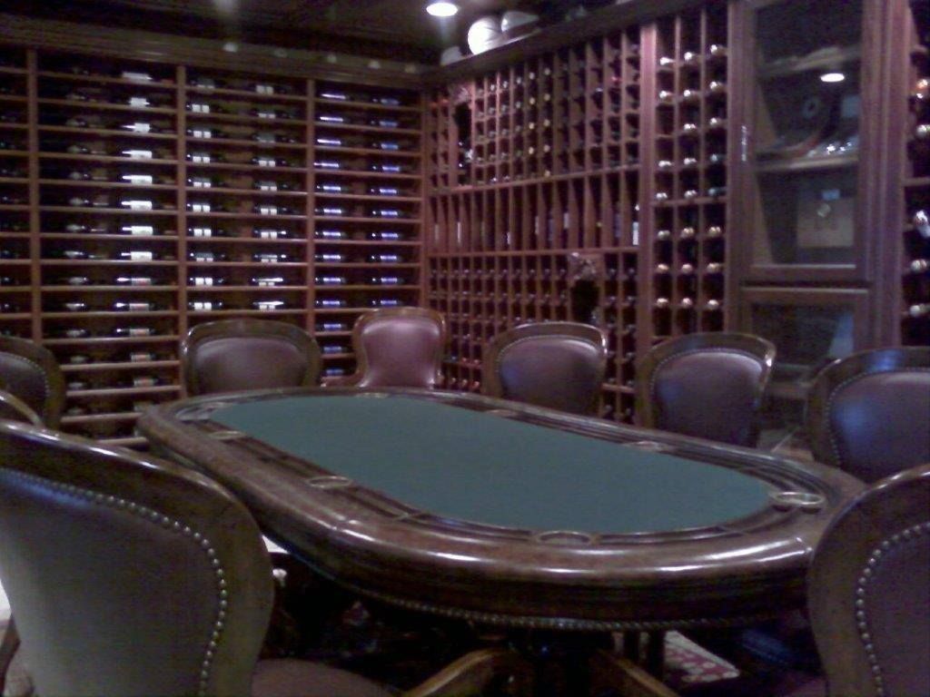 An oval poker table with green felt sits in a room with dark wood wine racks covering the walls.