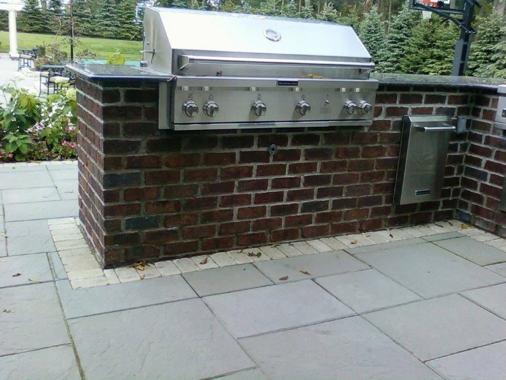 Built-in stainless steel outdoor grill set into a brick base on a stone patio in a backyard.