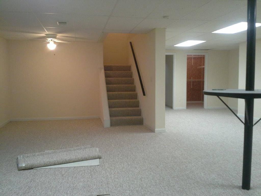 A carpeted, unfinished basement with carpeted stairs leading up, a black support pole, and a loose piece of carpet on floor.