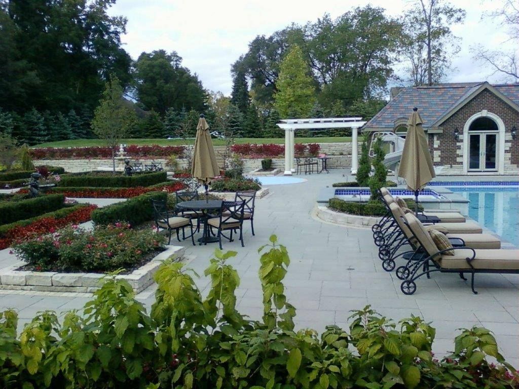 A backyard patio features a swimming pool, lounge chairs, a dining set, and structured landscaping with a stone pergola.
