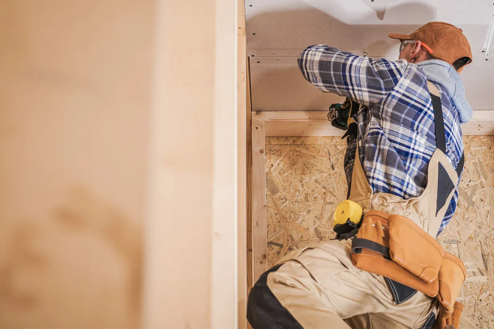A person in a plaid shirt and overalls works on a ceiling in a room with exposed wooden wall framing.