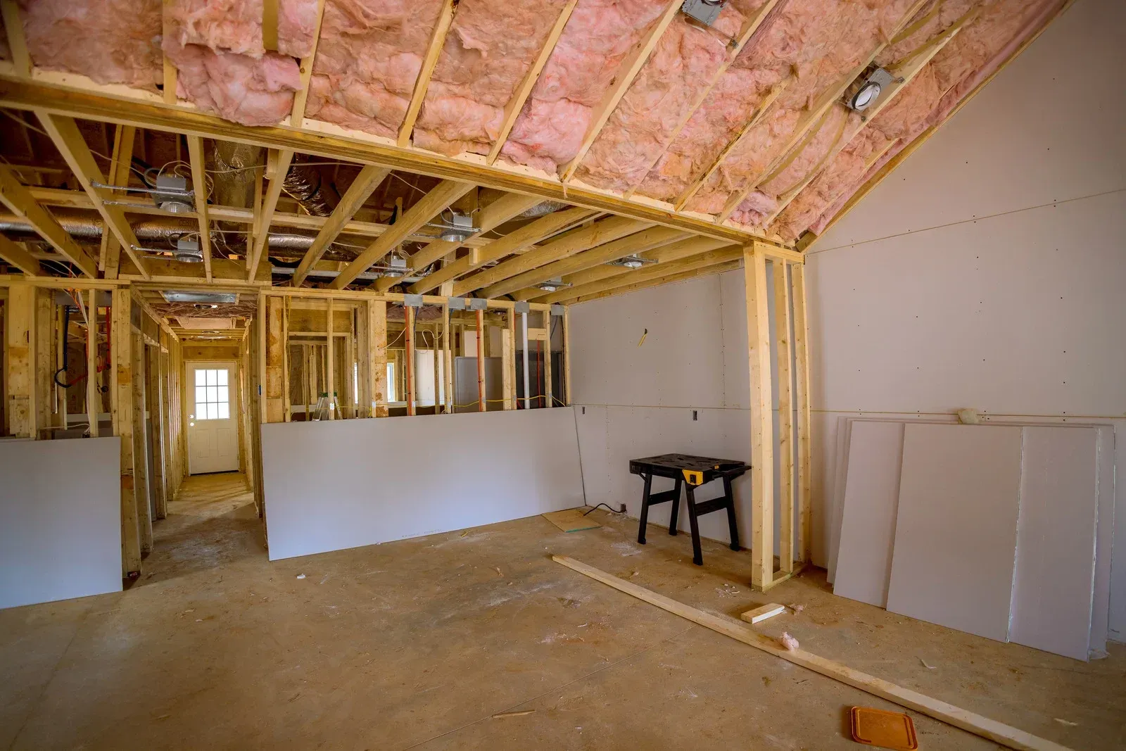 An unfinished room undergoing renovation, featuring exposed wooden beams, pink insulation, and drywall panels on the walls.