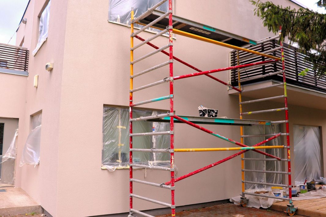 Tall metal scaffolding stands against the light pink exterior wall of a two-story house under renovation.