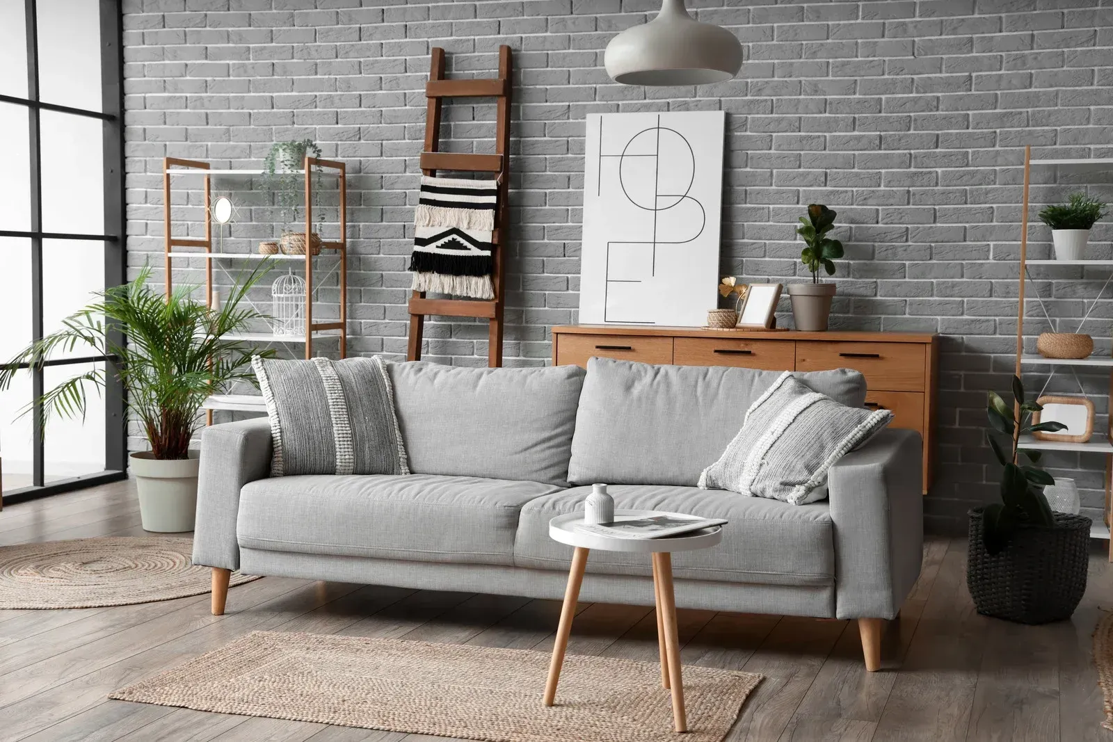 A light gray sofa sits against a gray brick wall, with a small coffee table, a wooden ladder, and indoor plants nearby.