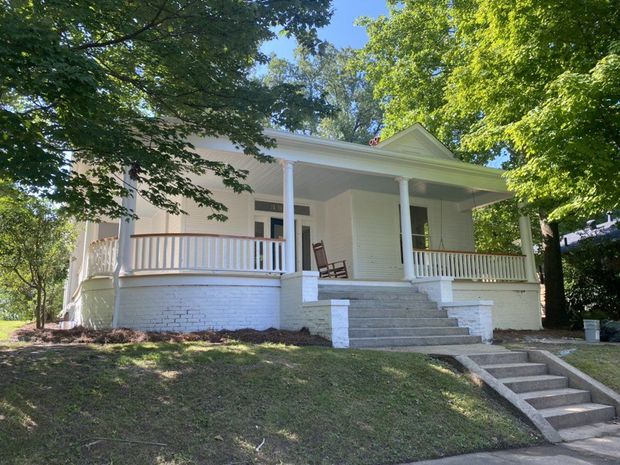 White house with porch, stairs, and trees on a sunny day.