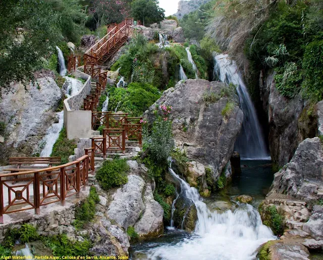 A waterfall is surrounded by rocks and trees and a wooden bridge.
