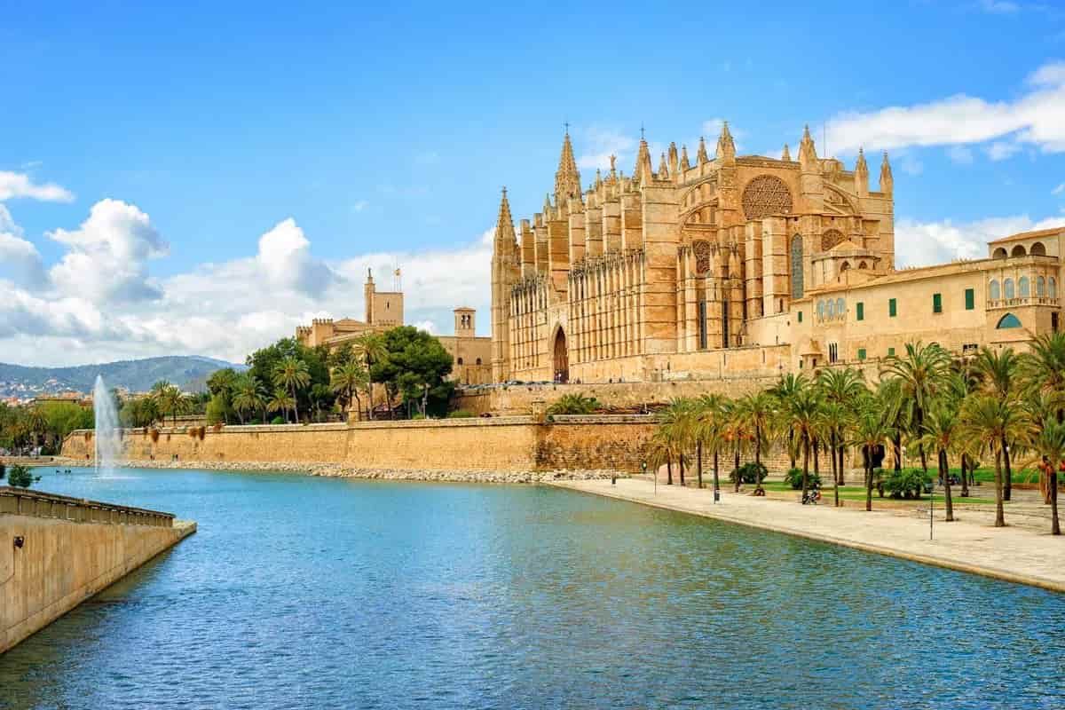 A large building in Palma de Mallorca with a fountain in front of it is next to a body of water.