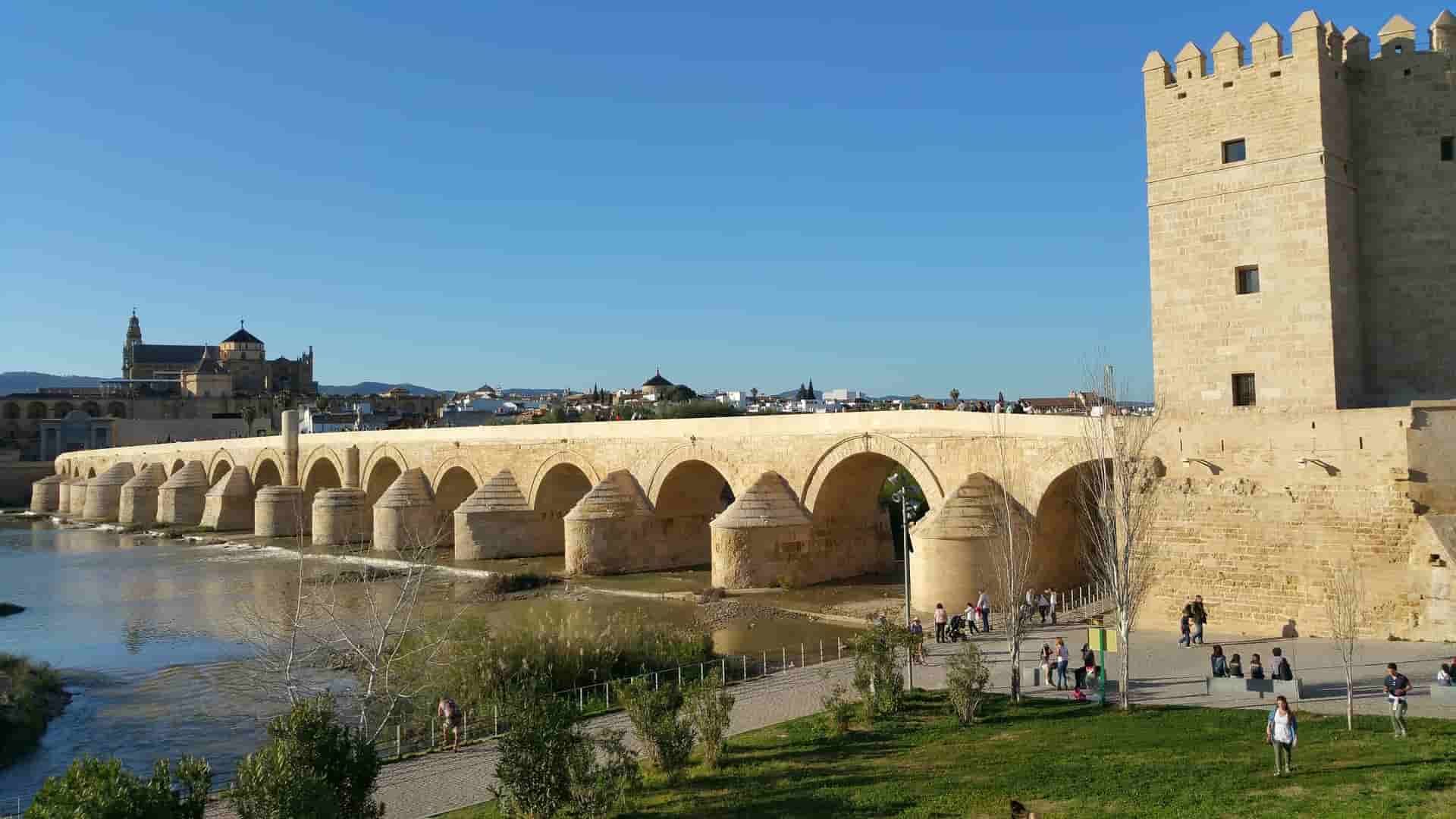 There is a bridge in Cordoba over a river in the foreground and a city in the background.