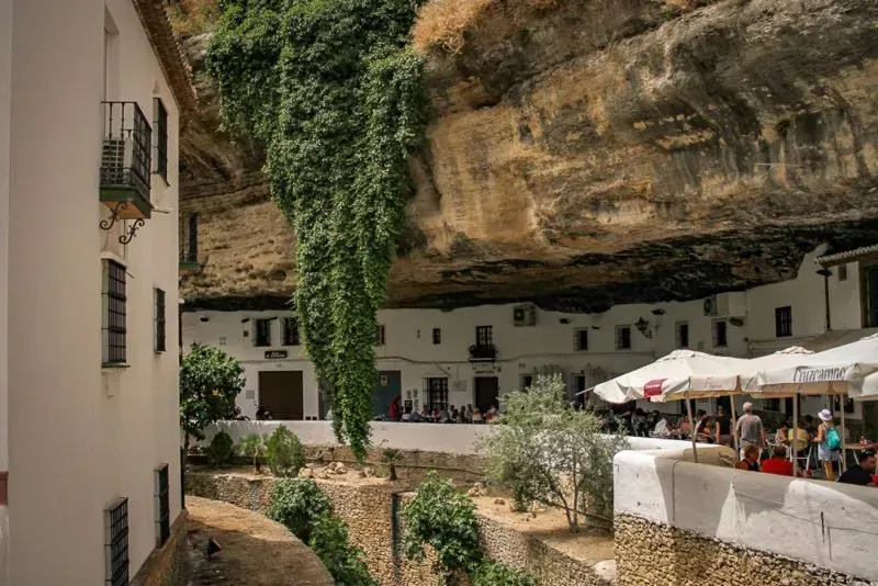 A group of people are sitting under umbrellas in front of a building in setenil.