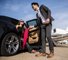 A person in a suit helps a passenger step out of a luxury car on an airport tarmac next to a private jet.