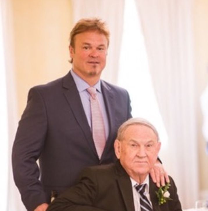 Man in suit with hand on older man's shoulder, both in a light-filled room.