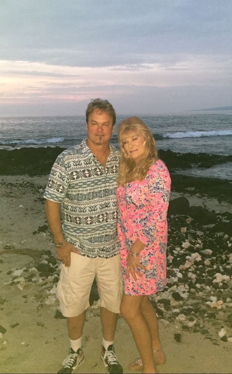 Man and woman on a beach at dusk. Man in shorts and patterned shirt, woman in pink patterned dress.