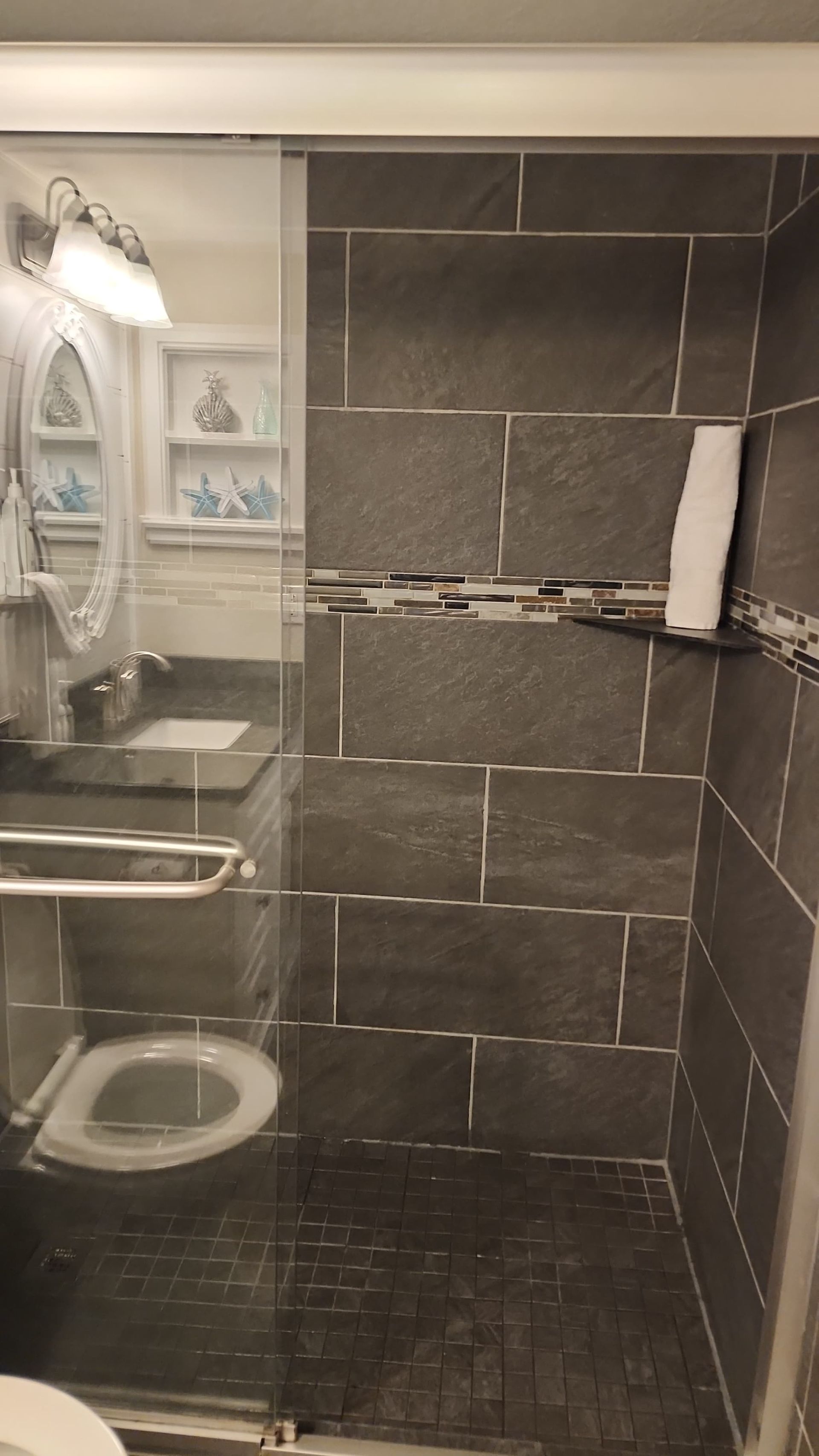 Shower with dark gray tile, glass door, white towel on a shelf, and a sink visible.