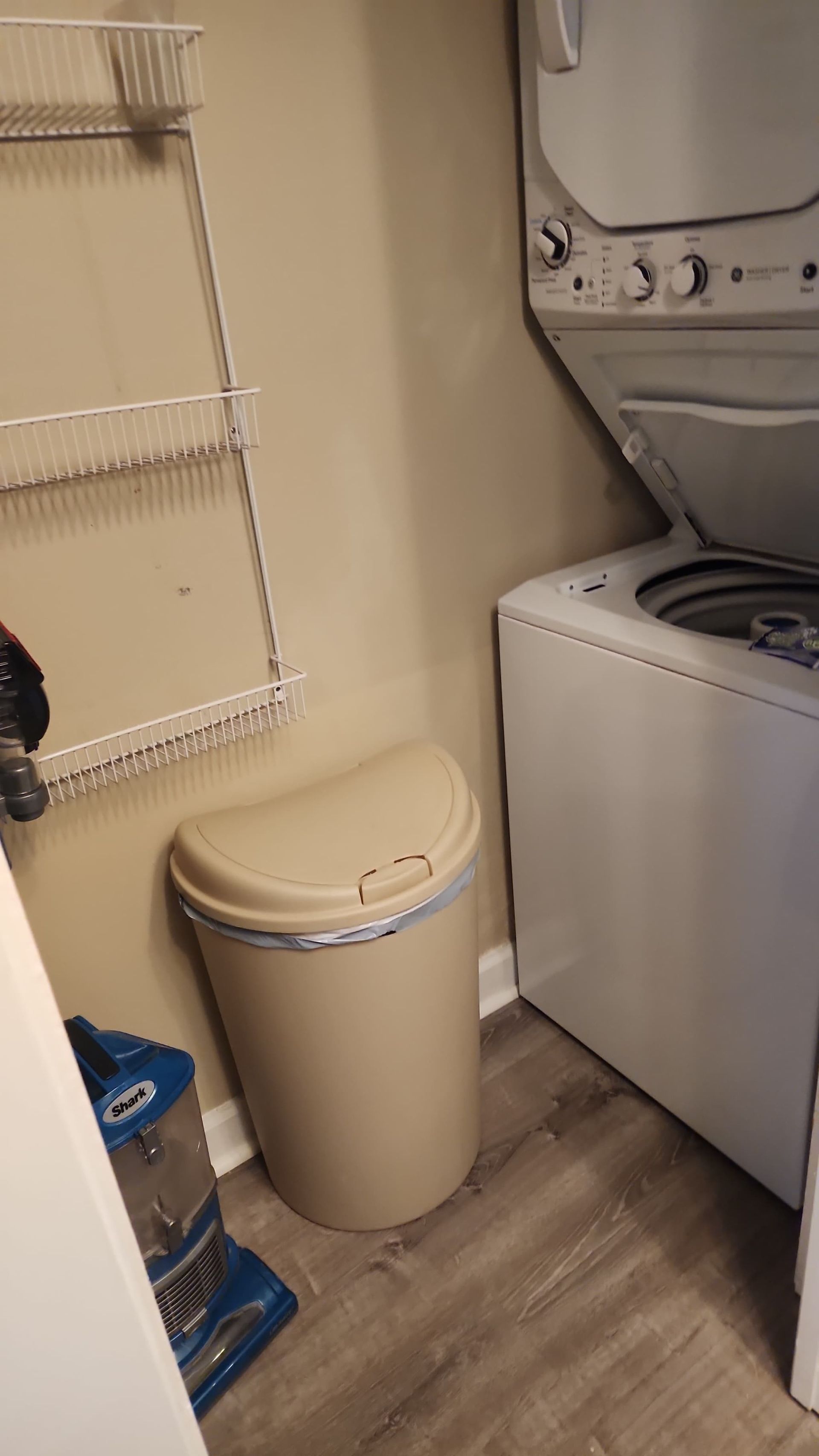 Laundry room with a stacked washer/dryer, a tan trash can, and a wall-mounted wire shelf.
