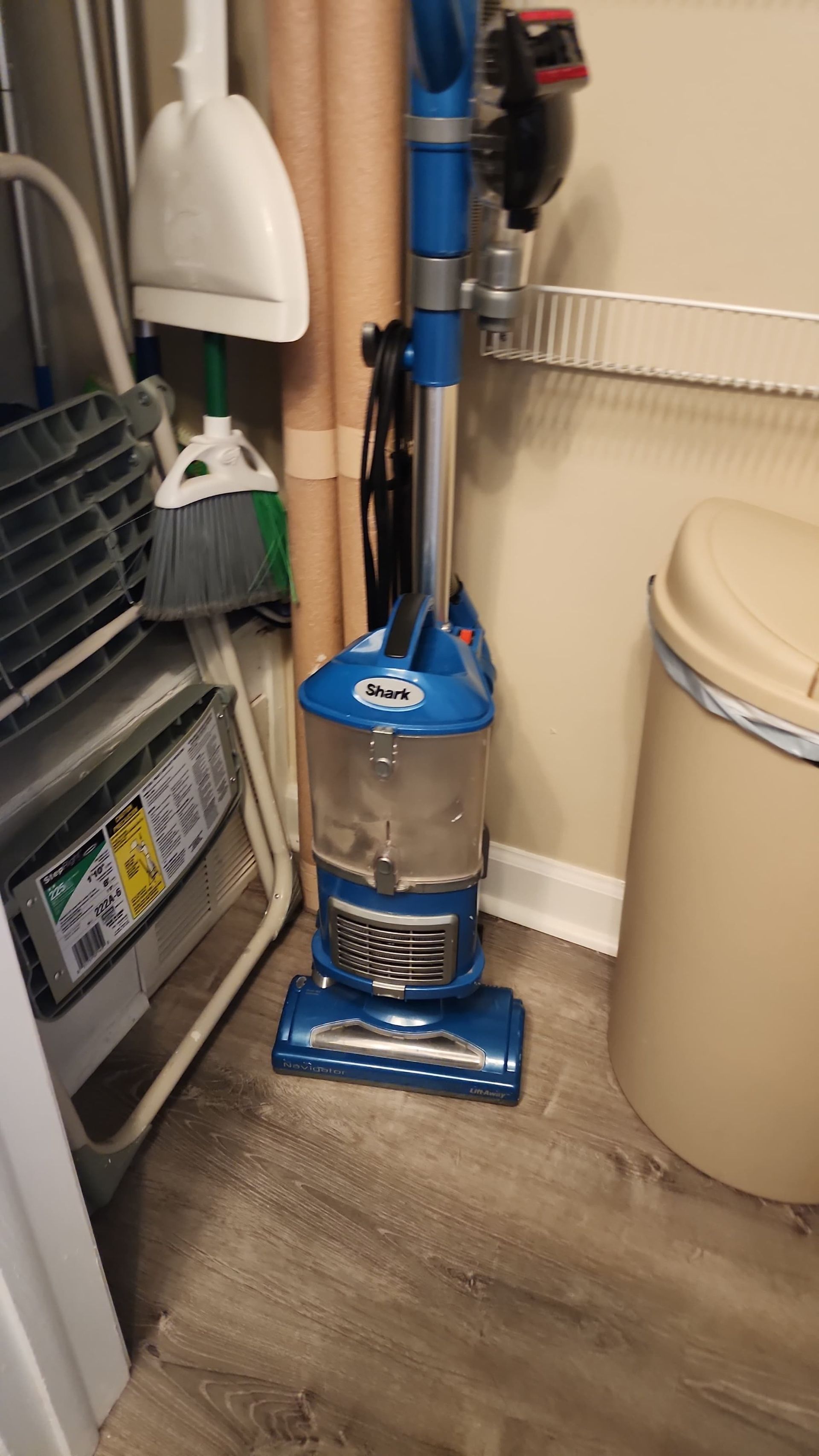 Blue upright vacuum cleaner in a utility room next to a trash can and cleaning supplies.