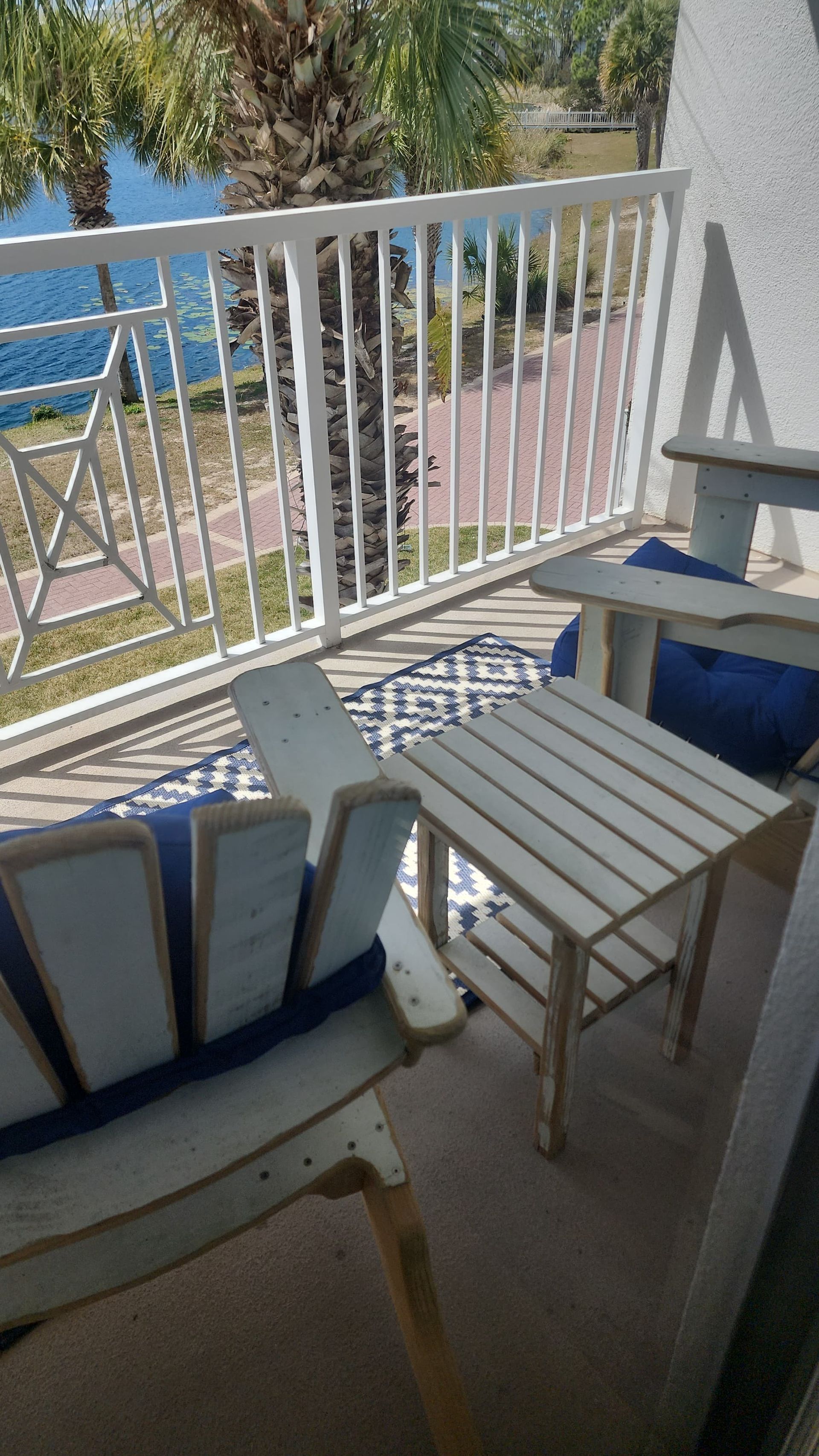 Balcony with Adirondack chairs, small table, blue cushions, patterned rug, and white railing overlooking water.