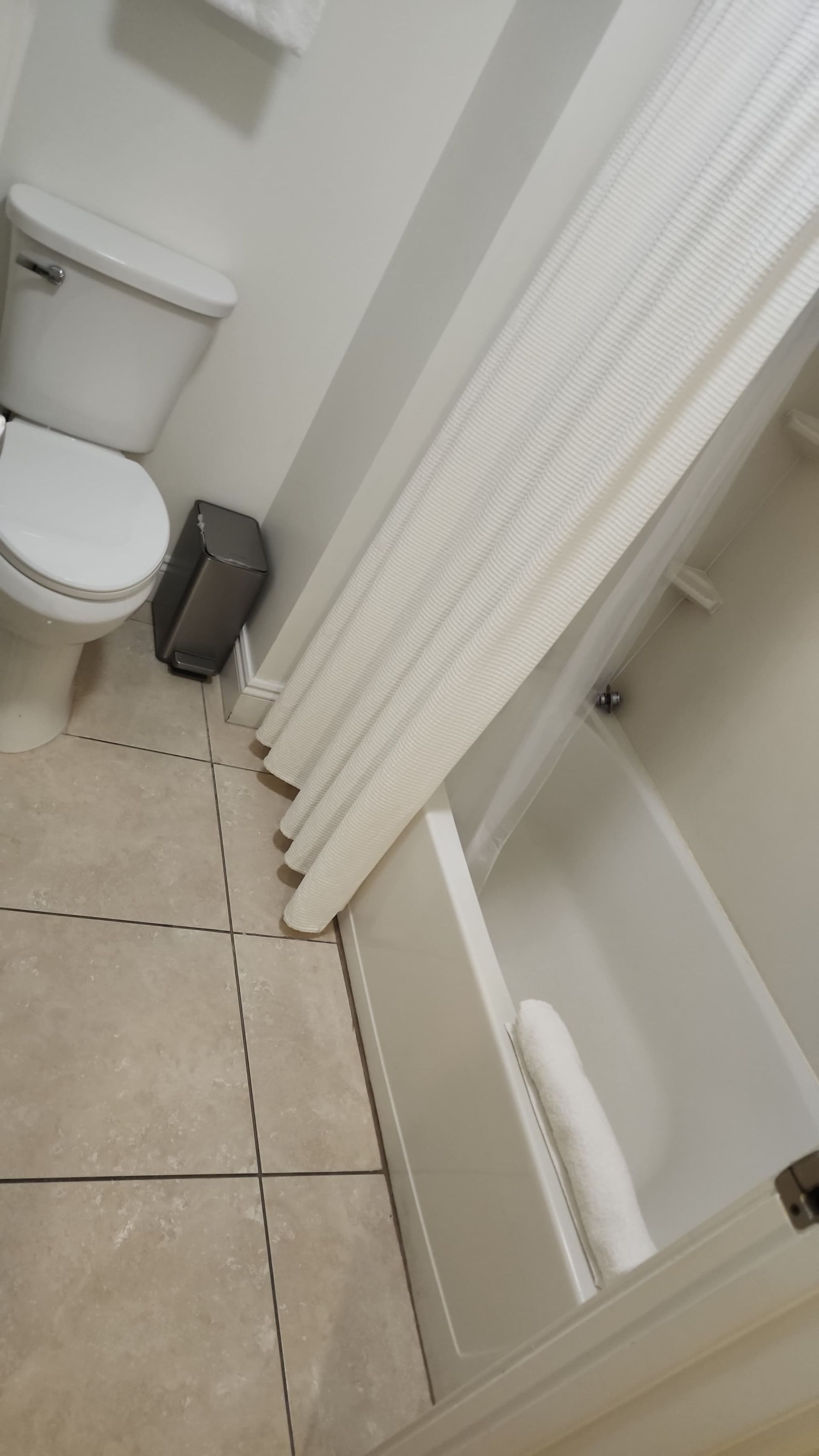 Bathroom with white tub, toilet, and curtain; tan tiled floor.