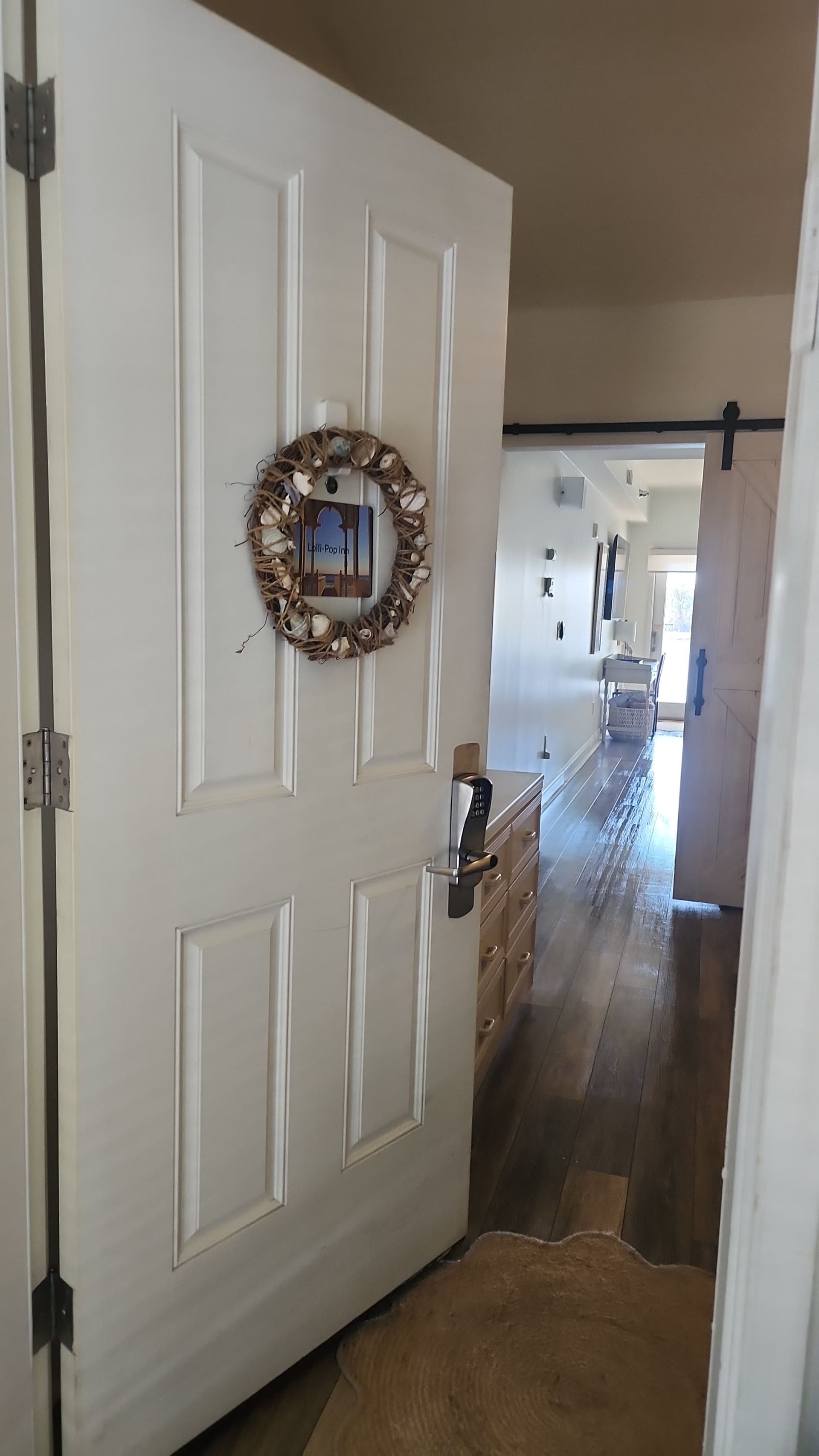 Open white door with seashell wreath, leads to a hallway with wood flooring and a partially visible kitchen.