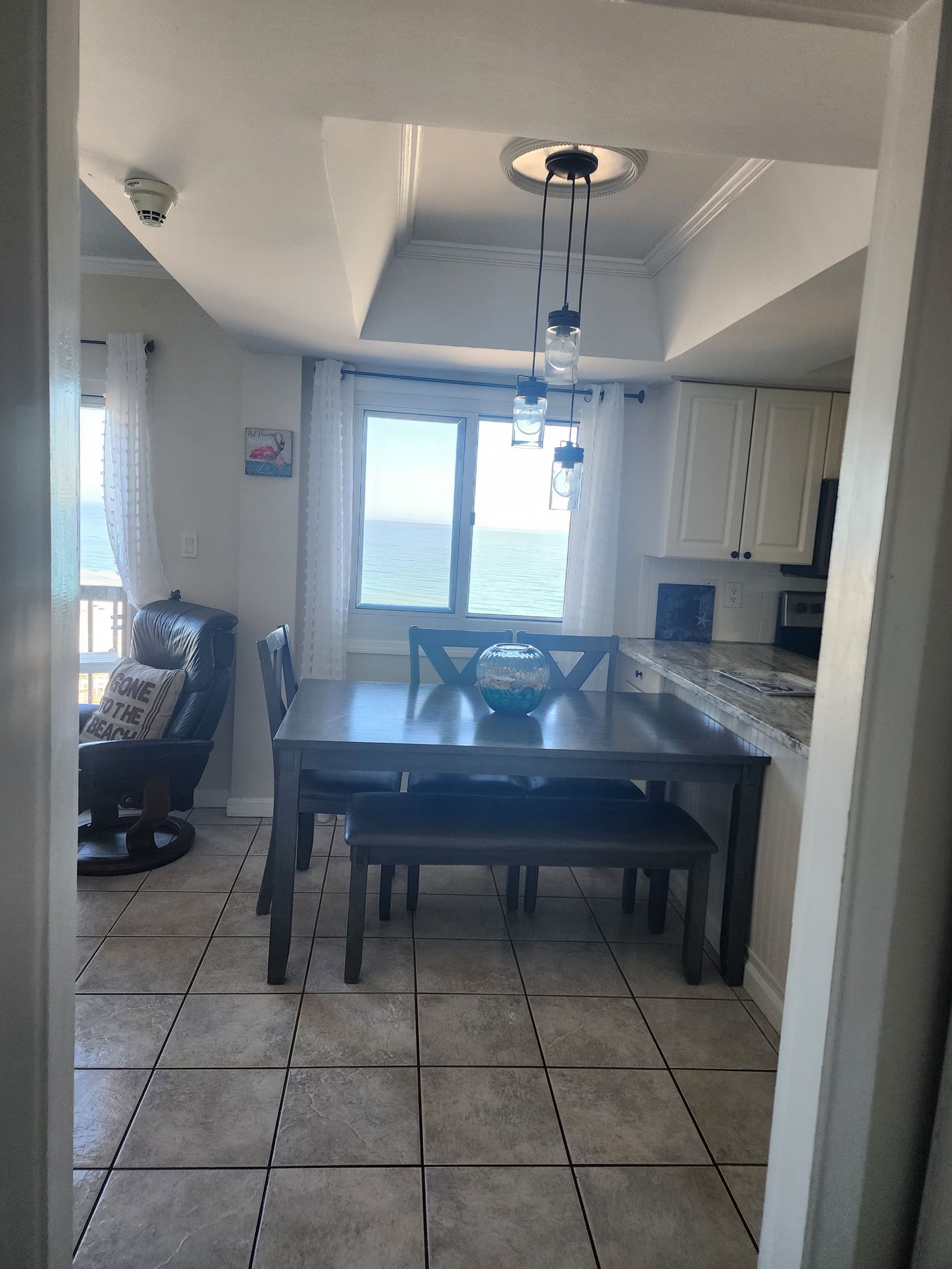 Dining room with a table, chairs, and a bench, overlooking the ocean through a window.
